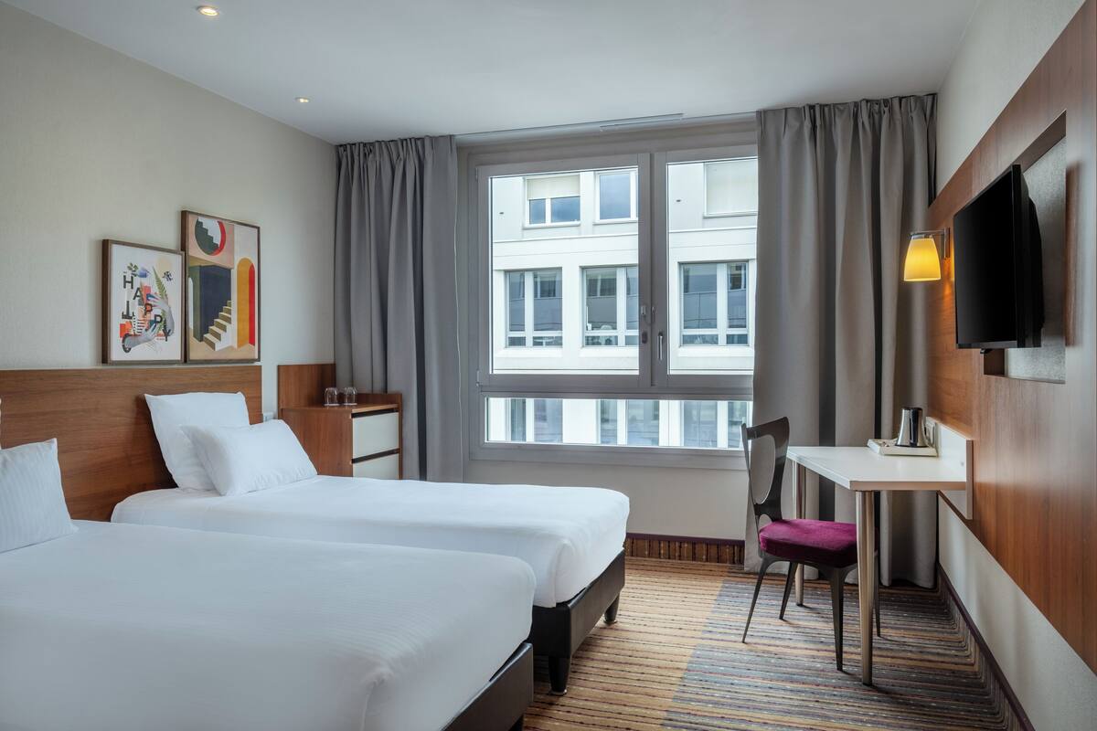 Ein Hotelzimmer mit zwei Betten, einem Schreibtisch, einem Sessel, einem Fernseher und einem Fenster mit Vorhängen.