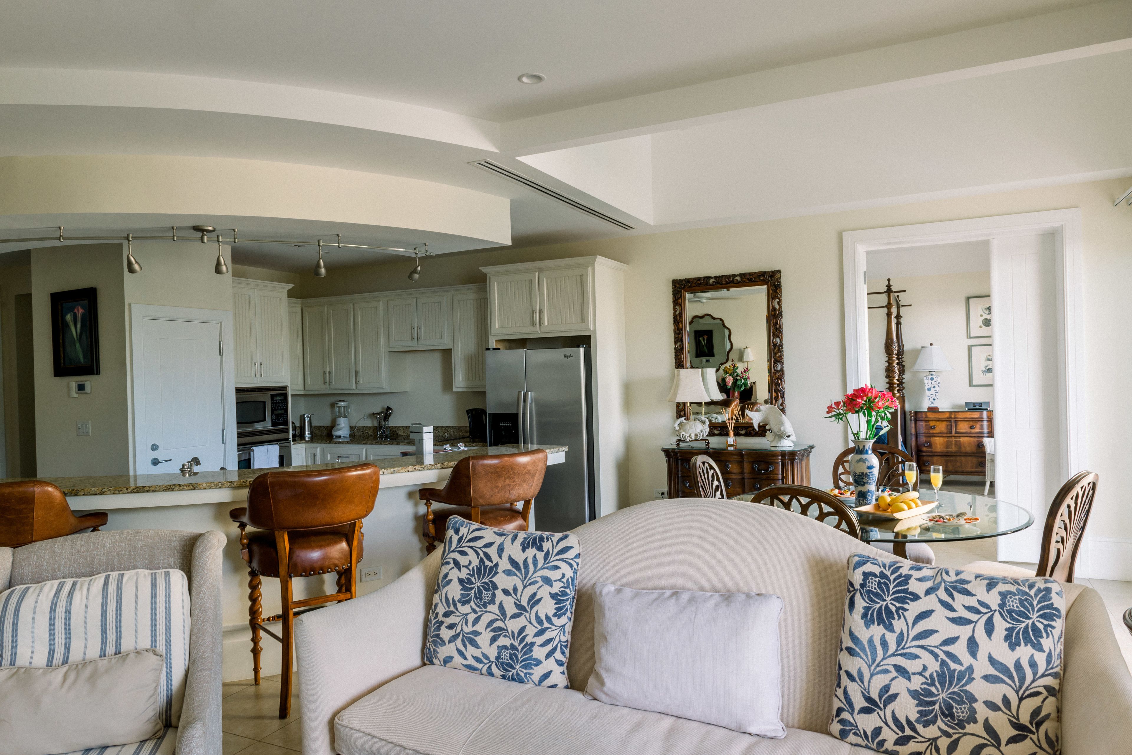 Un salón amplio con un sofá grande, una zona de comedor con una mesa de vidrio y una cocina con armarios blancos y un refrigerador.