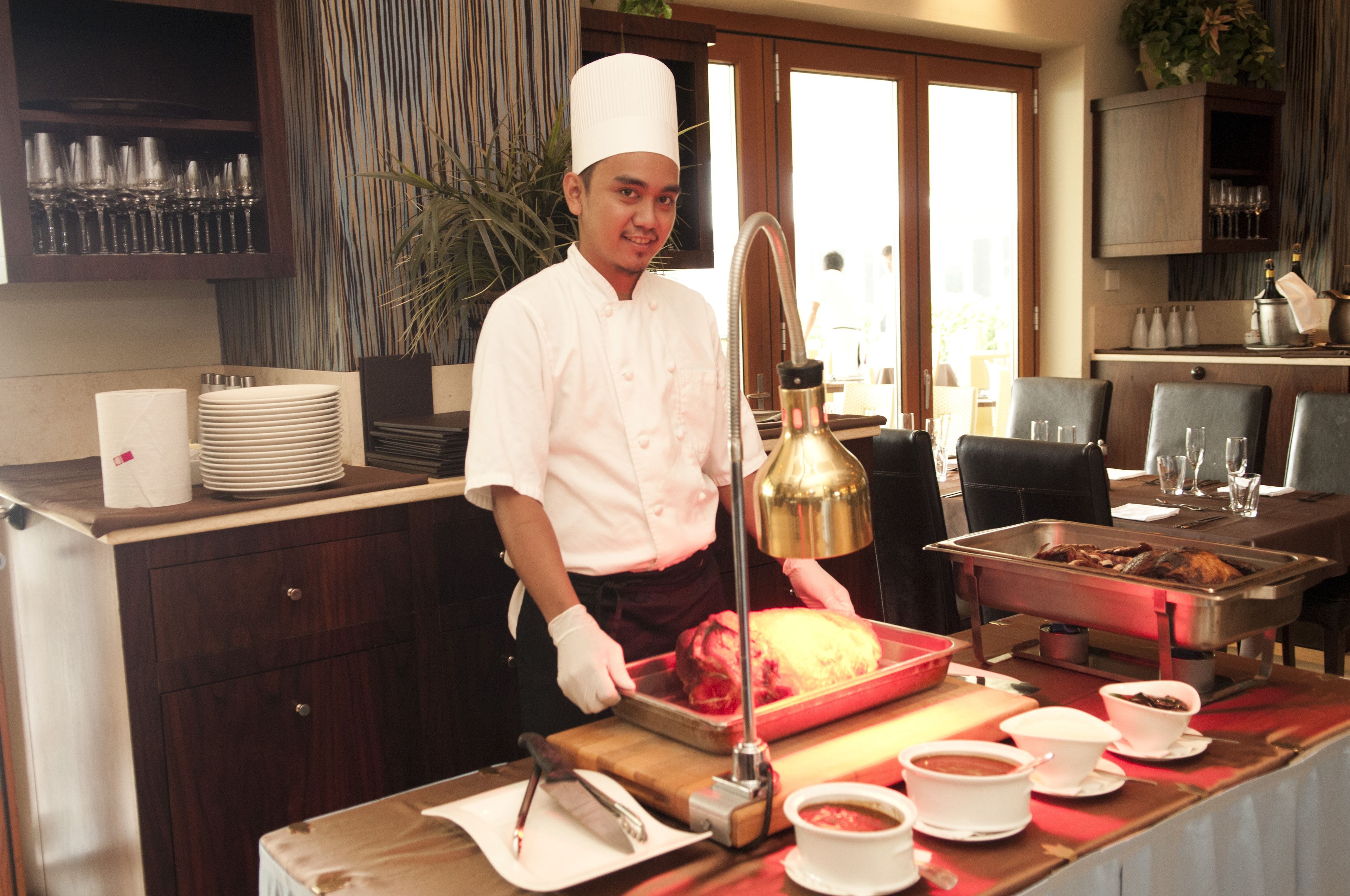 Un chef con uniforme blanco preparando comida en una cocina con mesas tipo buffet.
