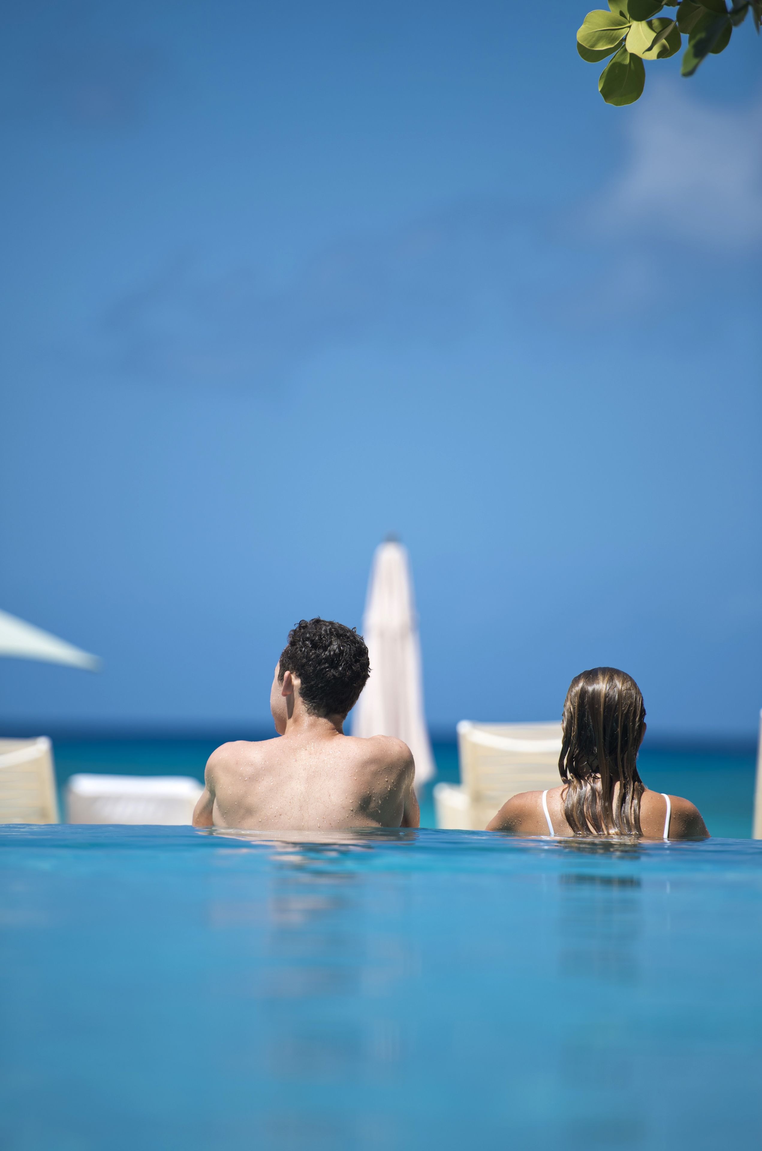 Un couple se détend dans une piscine à l’eau cristalline, avec vue sur l’océan et un parasol blanc en arrière-plan.