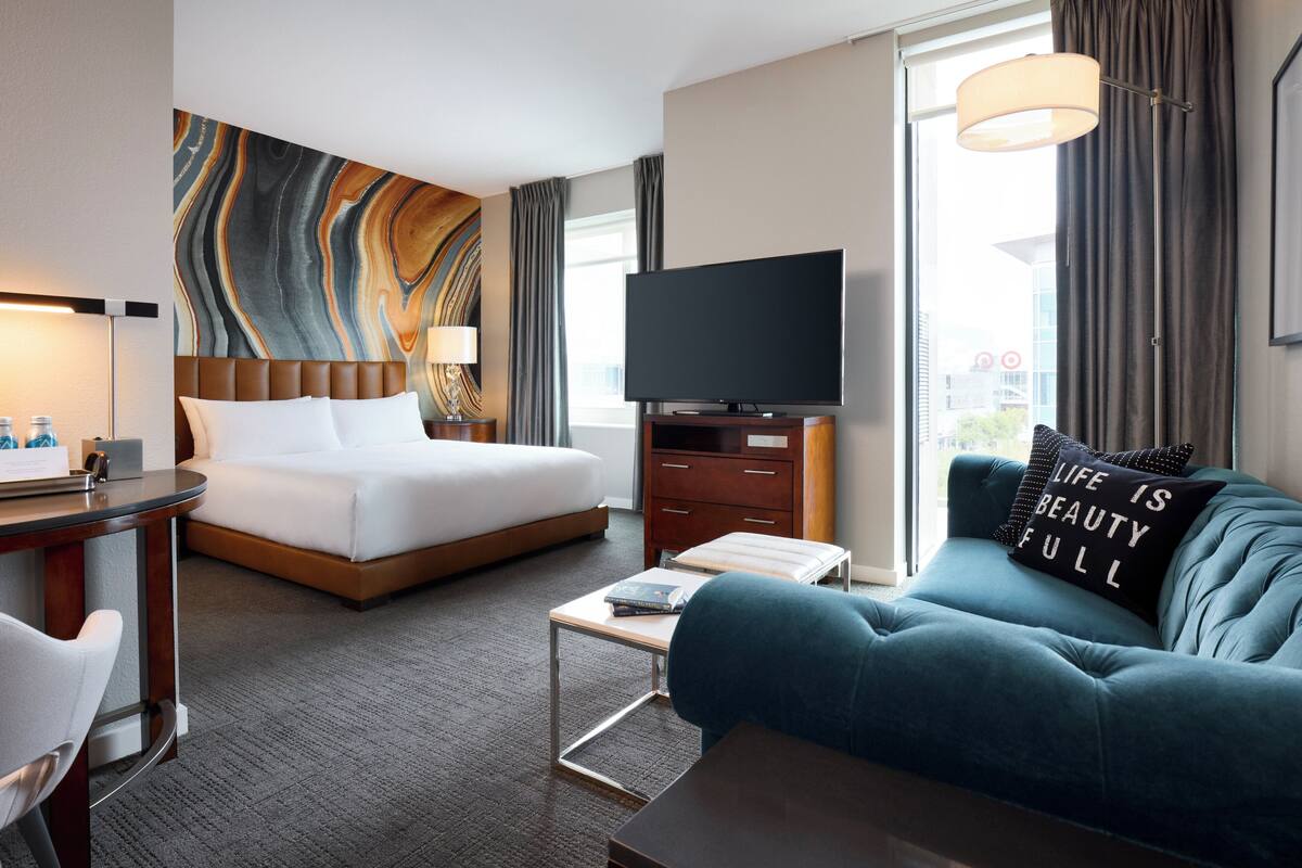 Habitación de hotel con cama, sofá, televisor, escritorio y ventana grande.