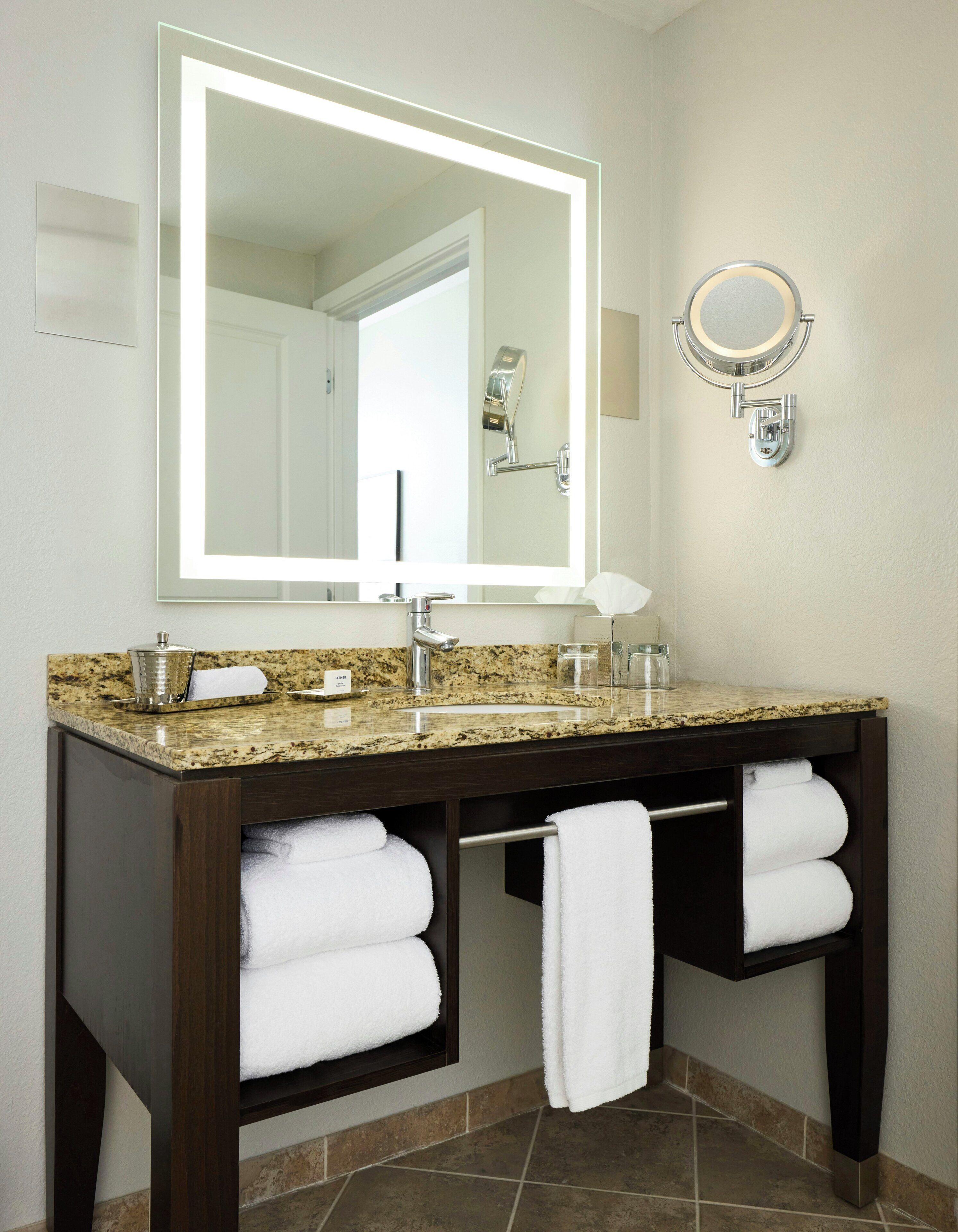 A bathroom with a granite countertop, a large mirror, a round vanity mirror, and two towels hanging on a rack.