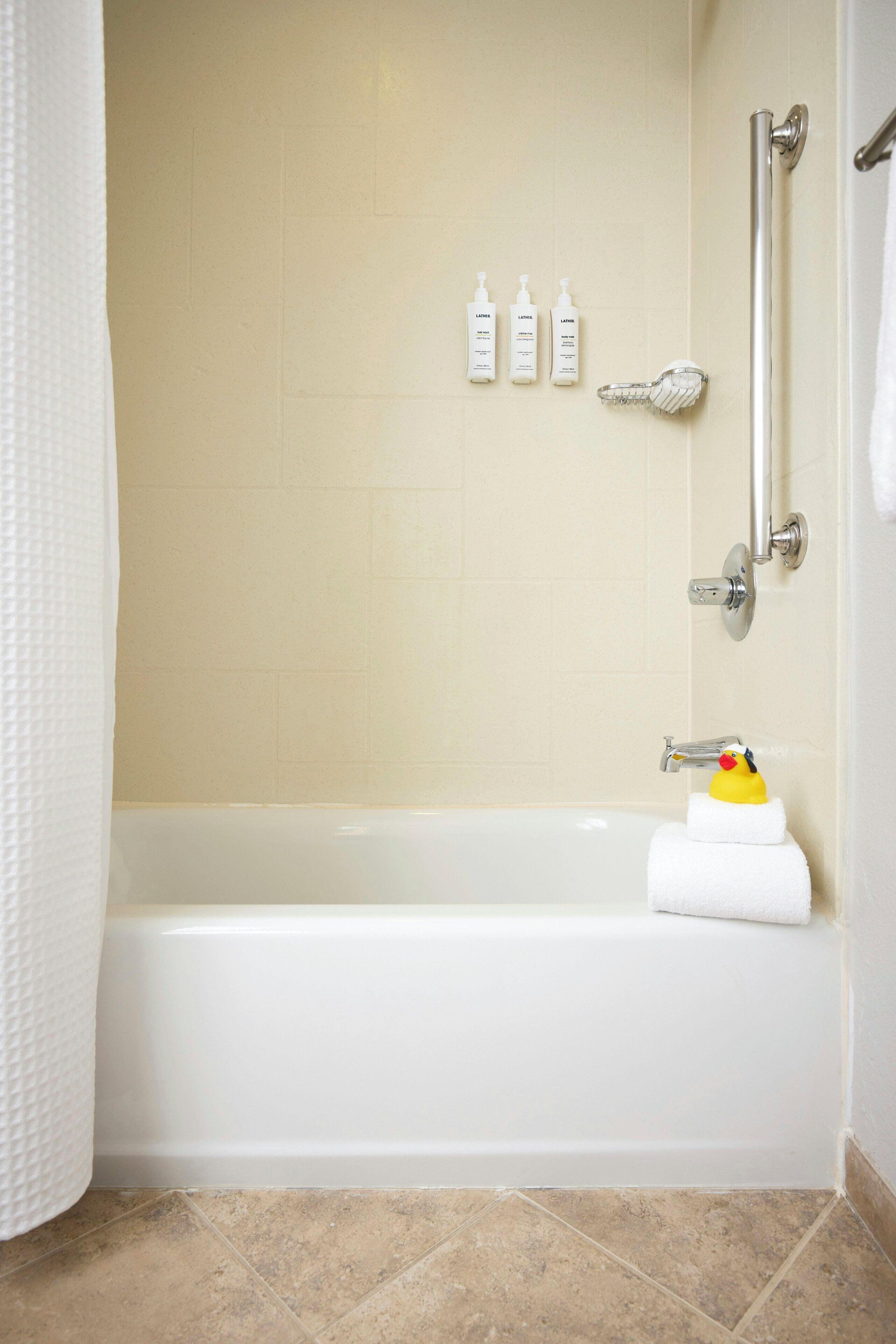 Un baño con bañera, ducha y barra de apoyo. Hay tres botellas de loción en la pared.