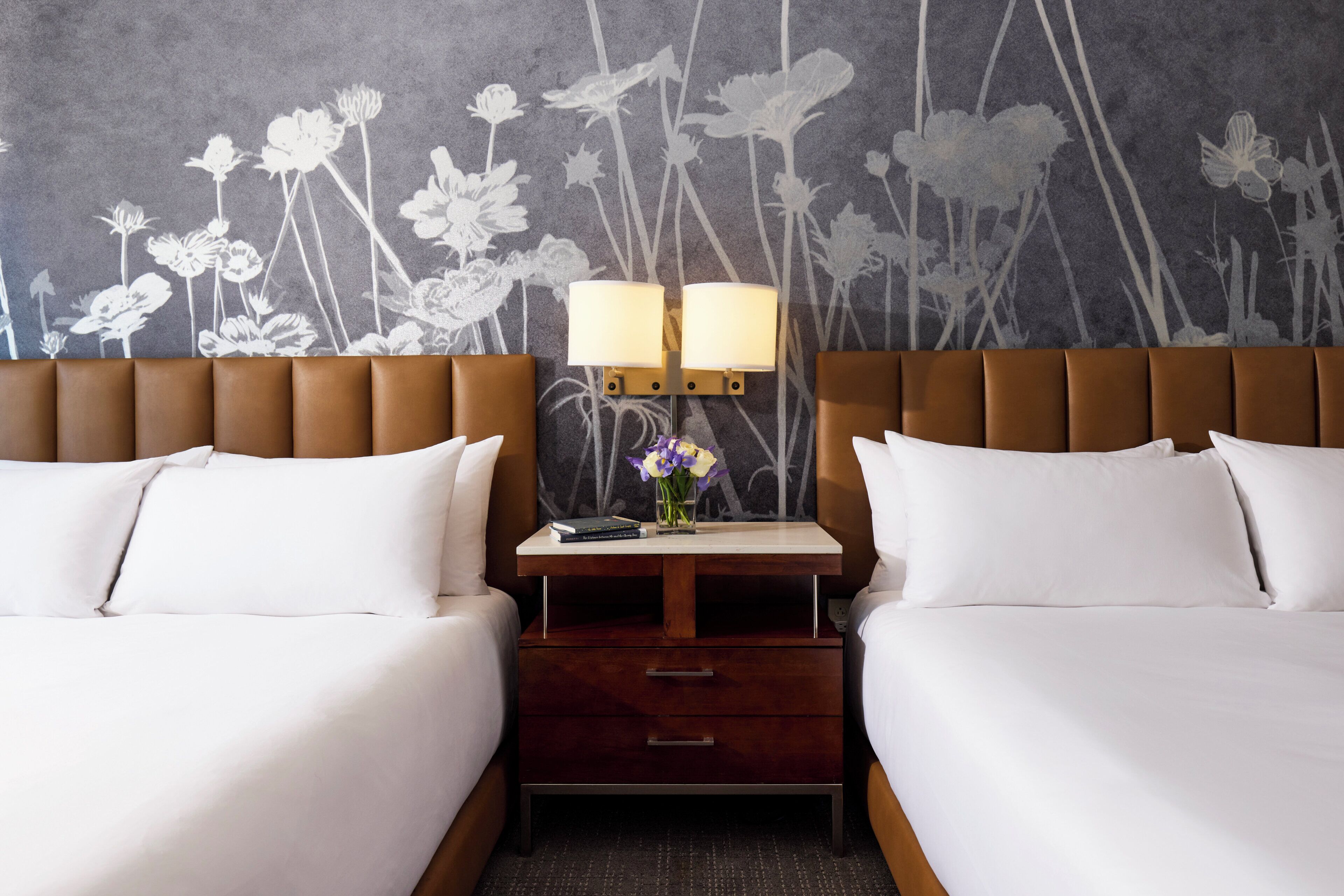 Ett hotellrum med två sängar, ett nattduksbord med en lampa och en väggmålning med blommotiv.