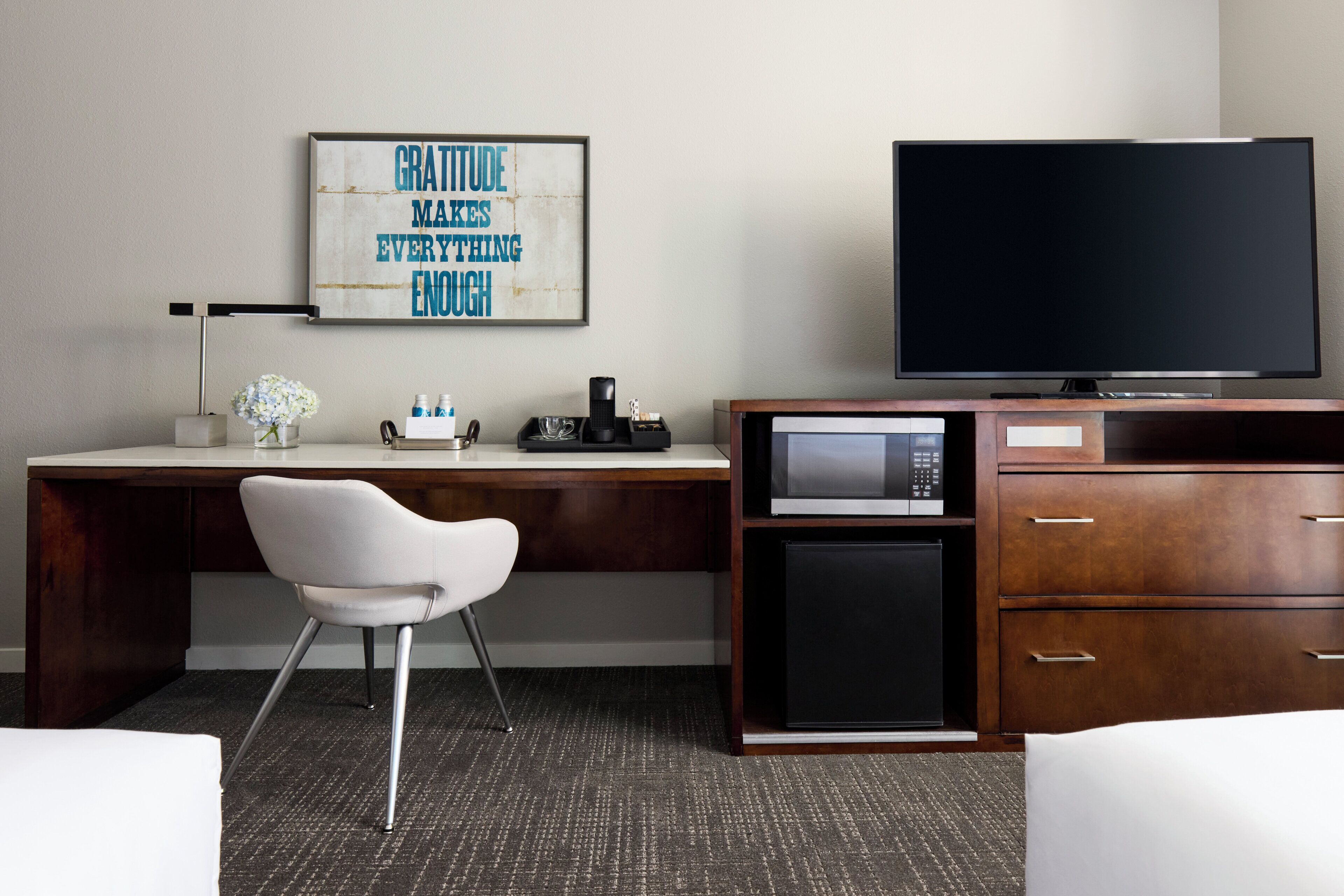 Habitación de hotel con un escritorio de madera, una silla blanca, un televisor de pantalla plana, un microondas y una cafetera.