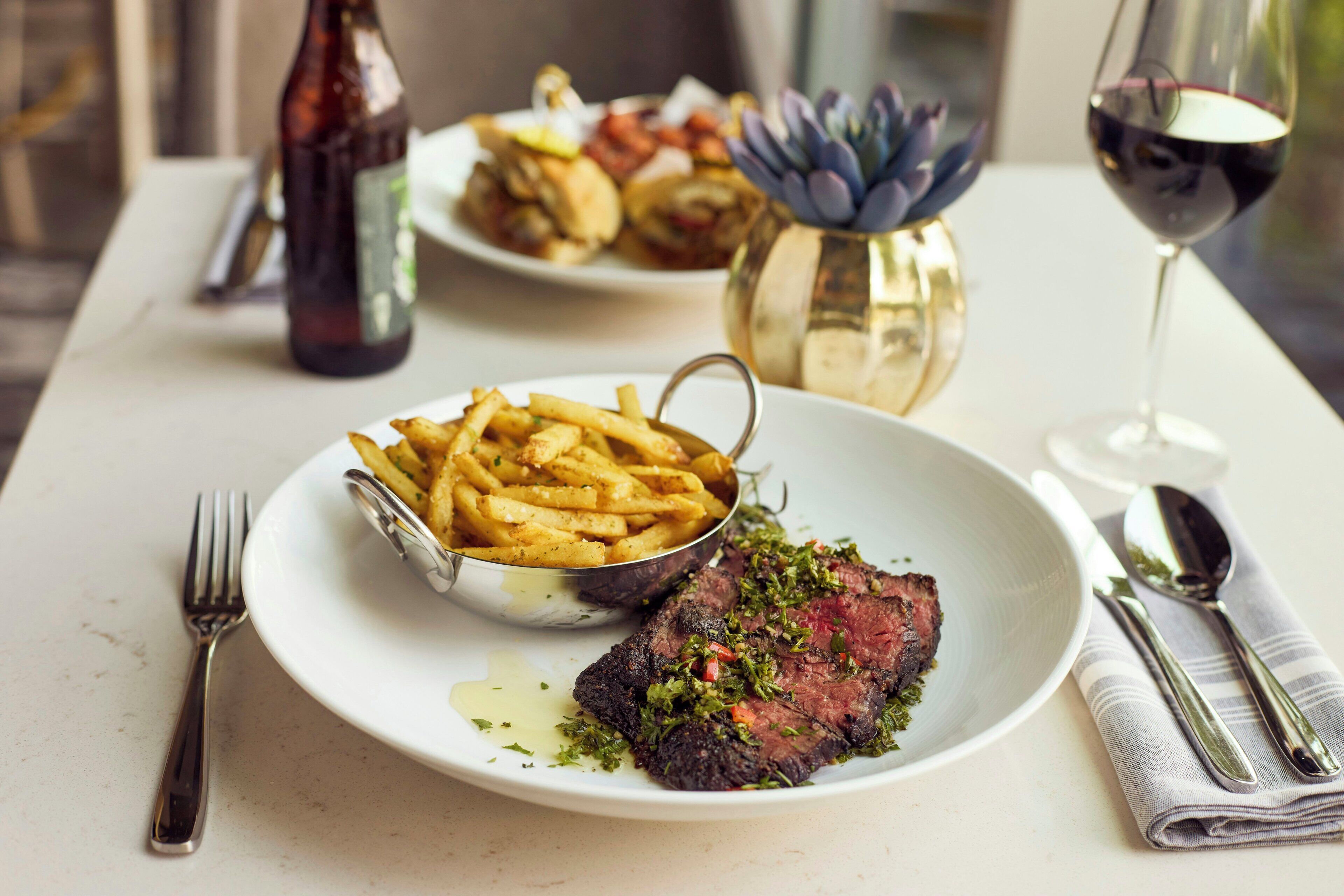 A dining table with a plate of steak and fries, a bottle, and a glass of red wine.