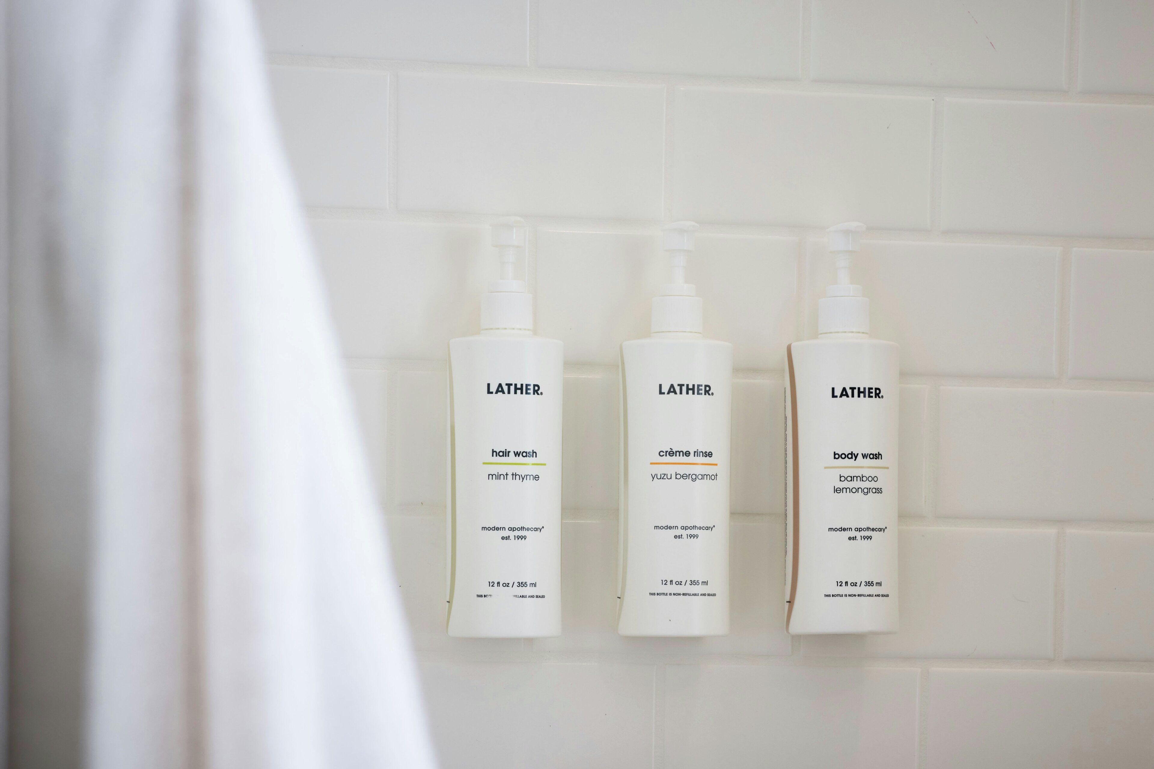 Three LATHER body wash bottles on a white tiled wall.