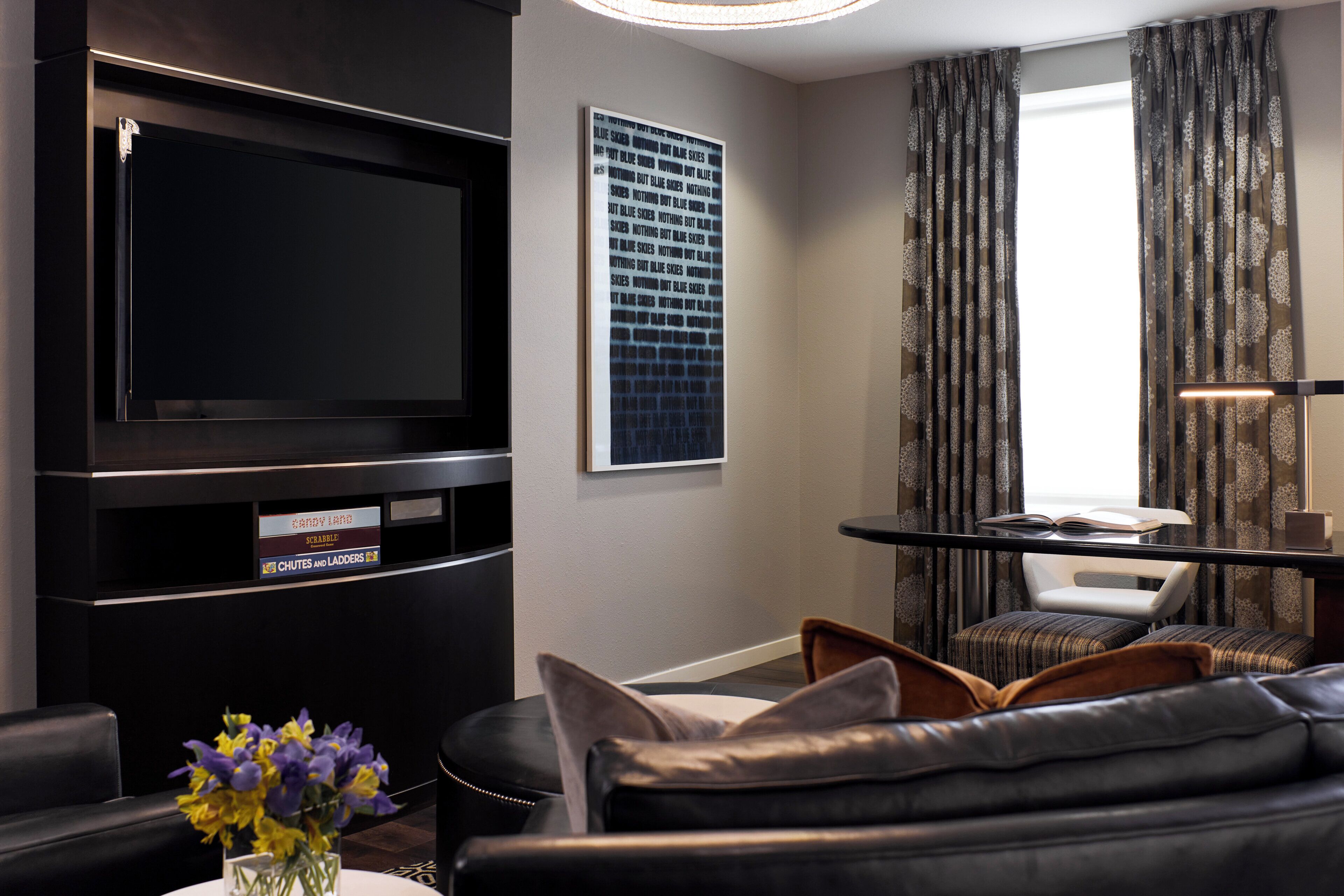 A modern living room with a black leather sofa, a flat-screen TV, a coffee table with flowers, and a large window with patterned curtains.