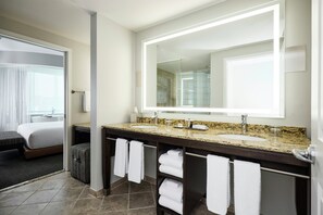 A hotel bathroom with a large mirror, a sink, and a shower. There is a bed visible in the adjacent room. - Archer Hotel Falls Church (Fairfax)
