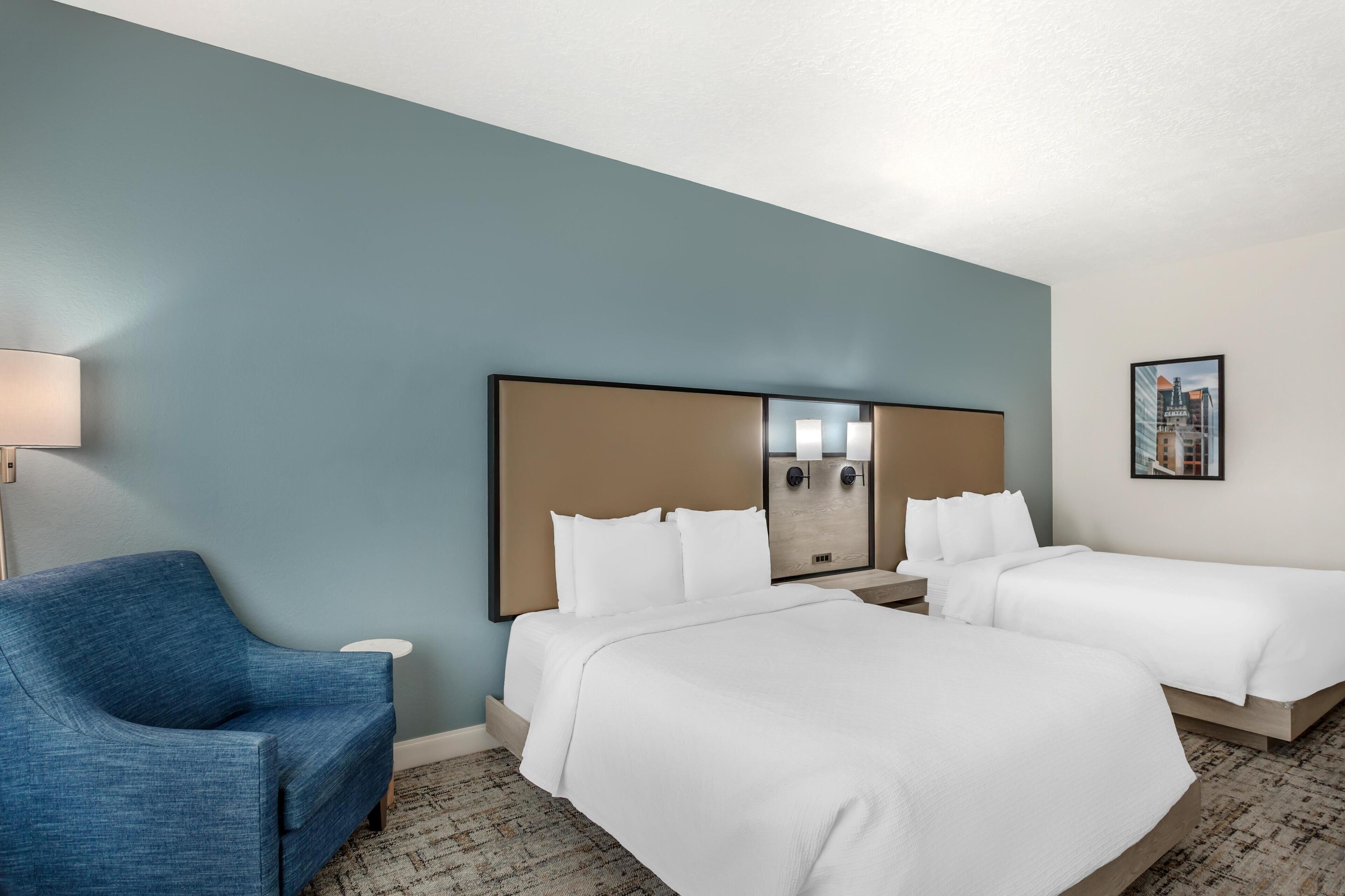 Habitación de hotel con dos camas, un sillón azul, una mesita de noche con una lámpara y un cuadro enmarcado en la pared.
