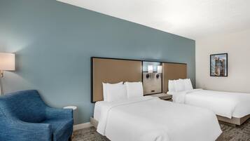 Habitación de hotel con dos camas, un sillón azul, una mesita de noche con una lámpara y un cuadro enmarcado en la pared.