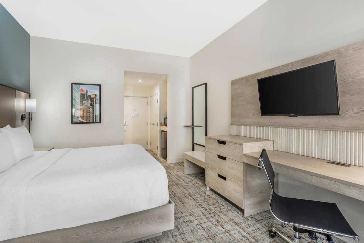 Une chambre d’hôtel moderne dotée d’un grand lit, d’un bureau avec une chaise, d’un téléviseur à écran plat fixé au mur et d’une œuvre d’art représentant un paysage urbain, également accrochée au mur.