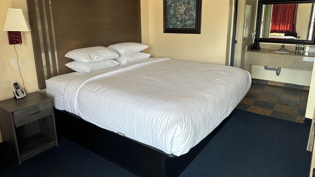 A hotel room with a large bed, a bedside table with a lamp, a telephone, a painting on the wall, and a bathroom with a sink and mirror. - Classic Inn Early (Early)