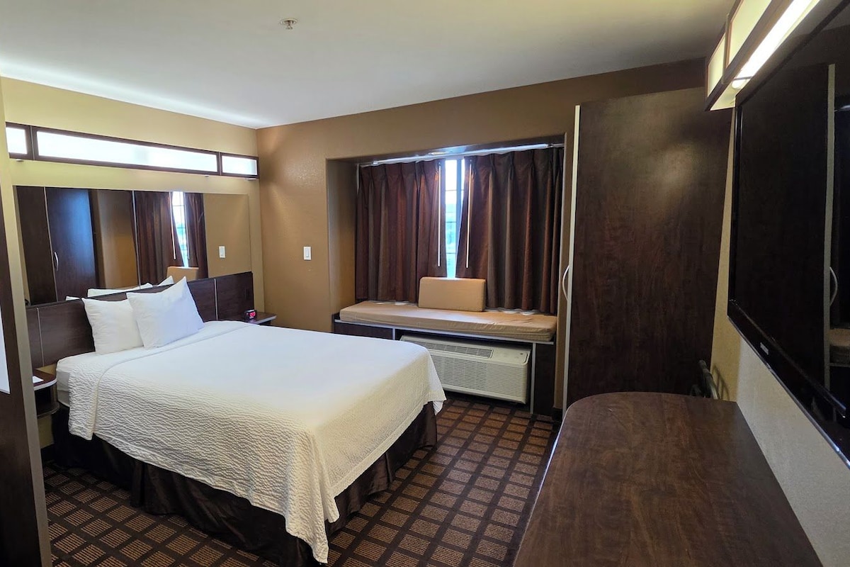 Une chambre d’hôtel comprenant un lit, un bureau, une chaise, une armoire et une fenêtre avec des rideaux.