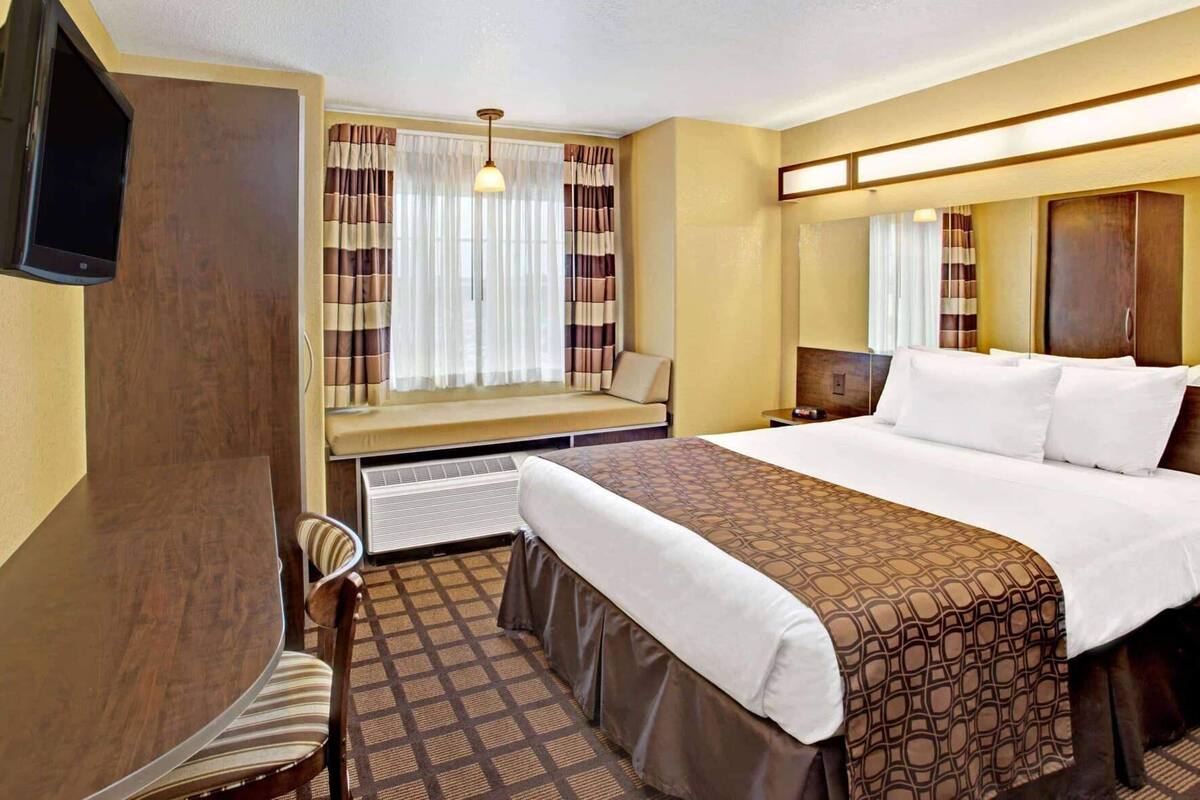Ein Hotelzimmer mit einem Bett, einem Schreibtisch, einem Stuhl, einem Fernseher und einem Vorhangfenster.