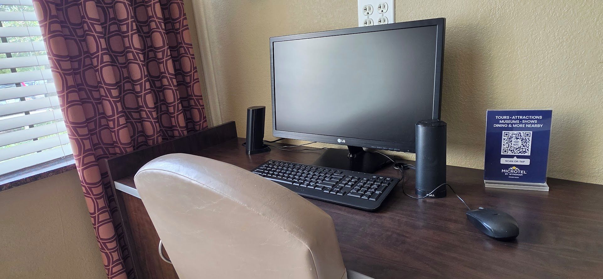 Una camera d'hotel con una scrivania, un computer, una sedia, un mouse e un cartello con informazioni su tour e attrazioni.