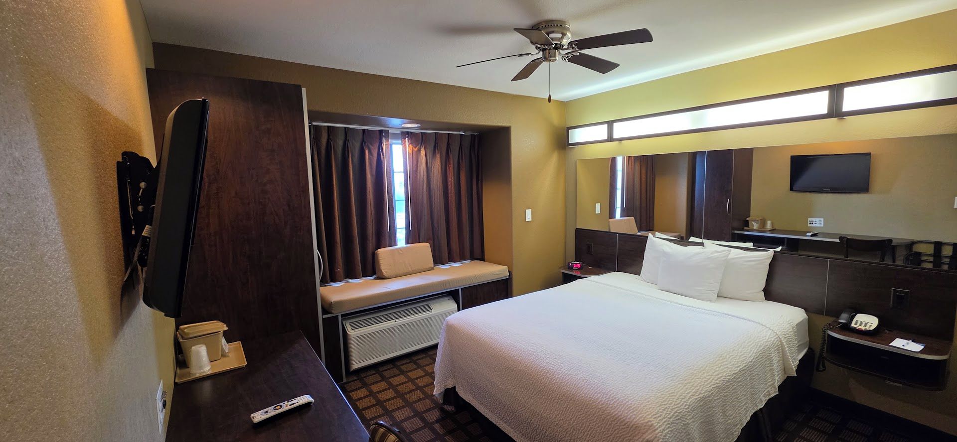 Habitación de hotel con cama, escritorio, silla, televisor, ventilador de techo y ventana con cortinas.