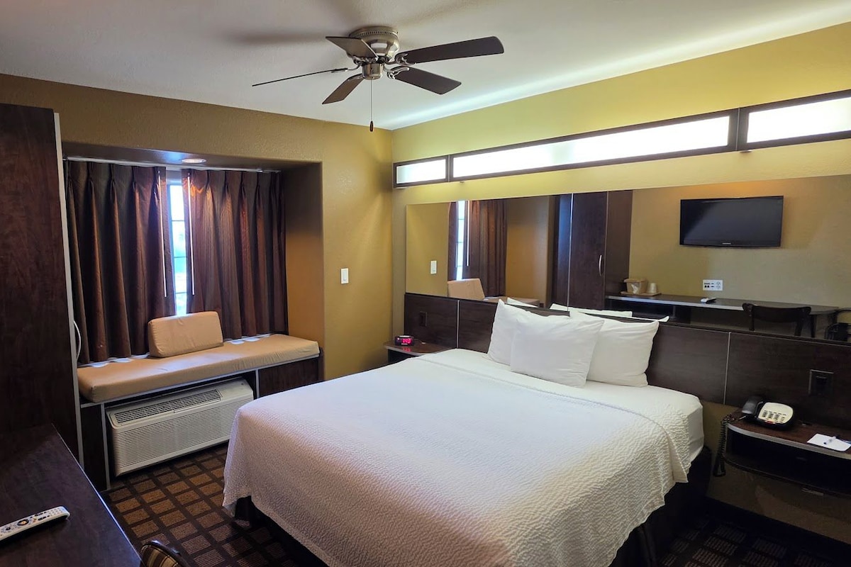 Une chambre d’hôtel équipée d’un lit, d’un bureau, d’une chaise, d’une télévision, d’un ventilateur de plafond et d’une fenêtre avec des rideaux.