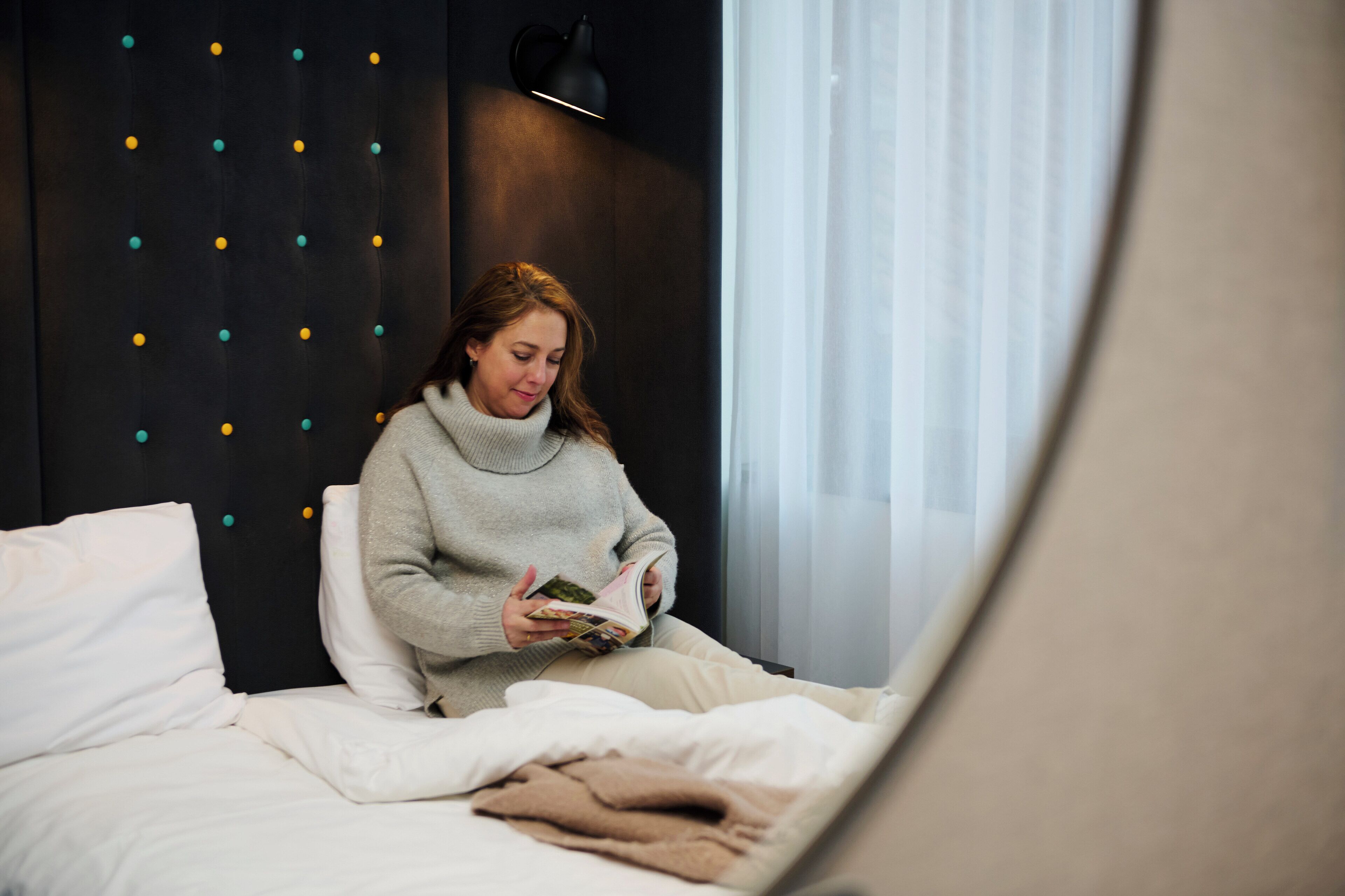 A person sitting on a bed in a room with a dark headboard, reading a book.