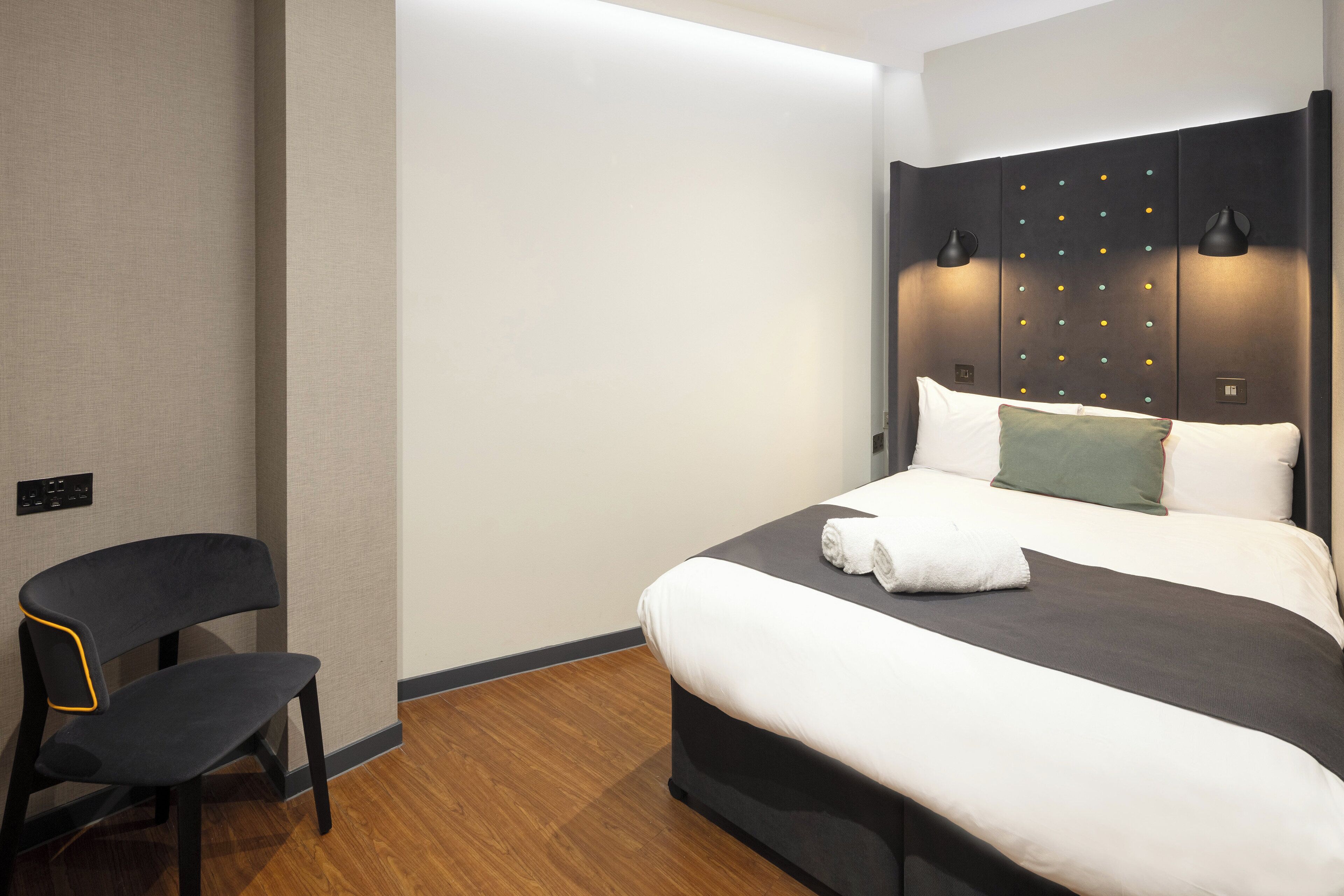A modern hotel room with a large bed, a chair, and a headboard with lights.