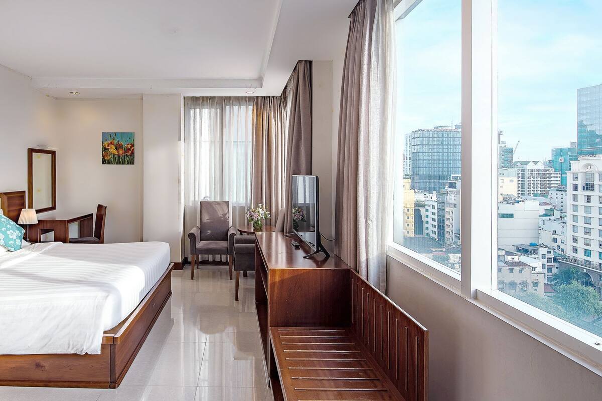 A hotel room with a bed, desk, chair, and a large window with a city view.