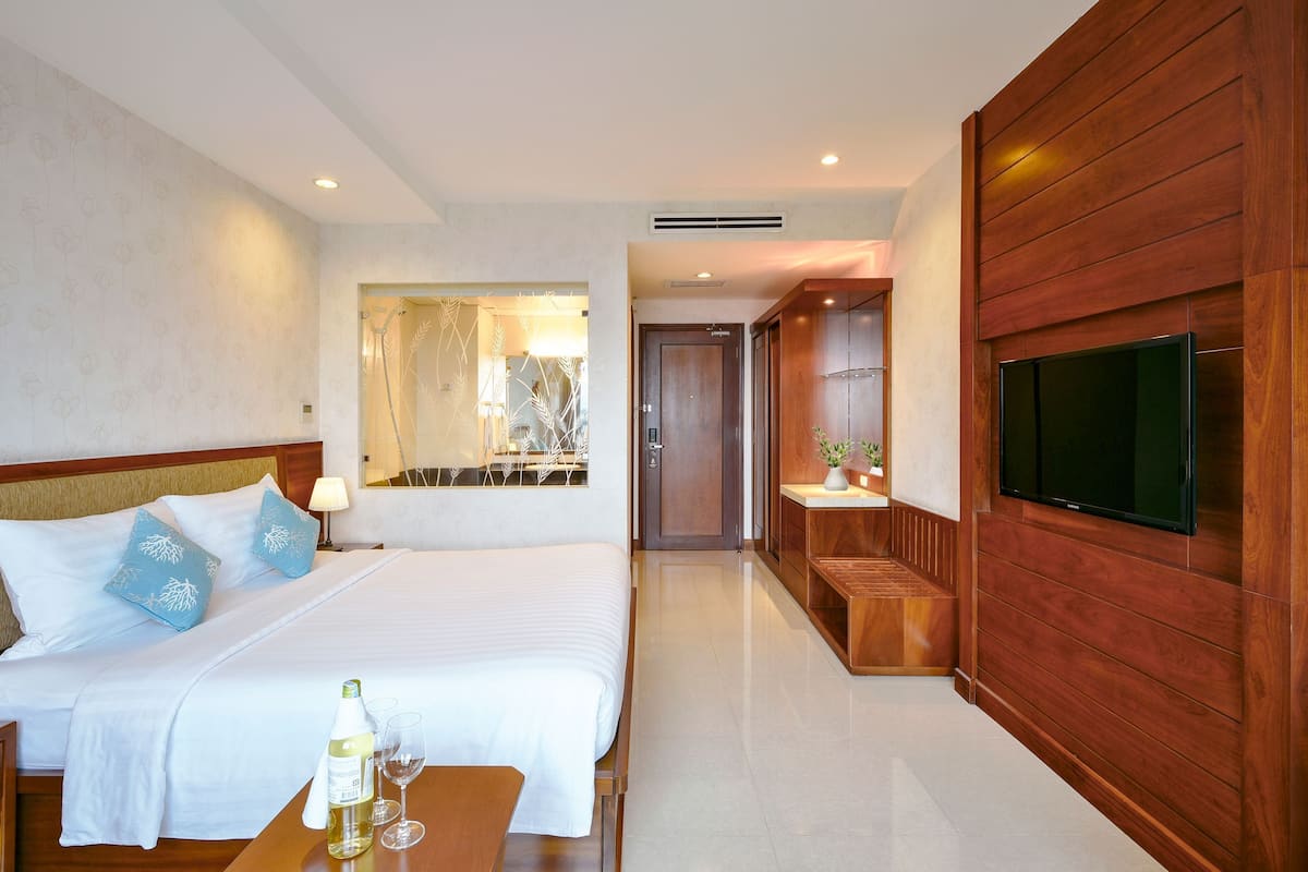 A hotel room with a large bed, a wooden wall-mounted TV, a bedside table with a bottle and glasses, and a mirror with a decorative frame.