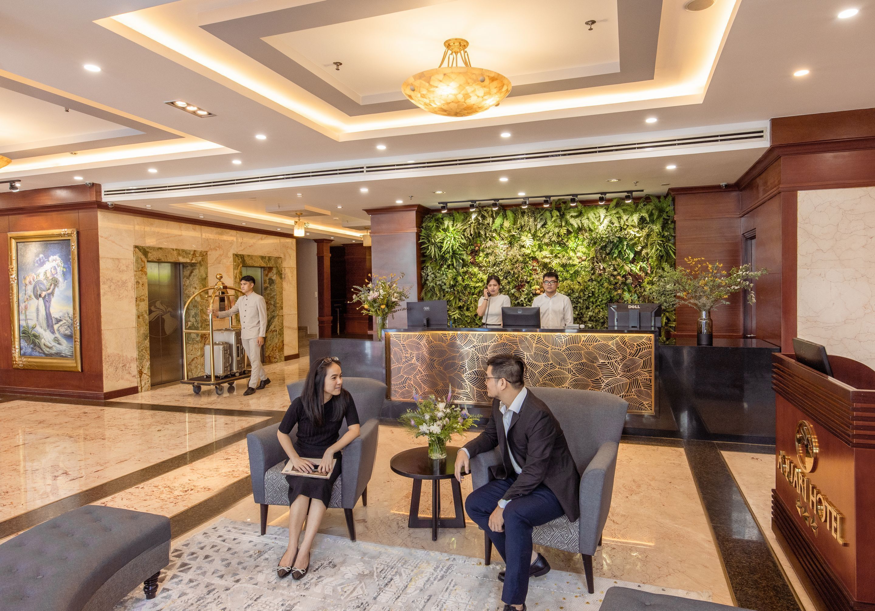 Vestíbulo de hotel con una recepción, dos huéspedes sentados y un hombre trajeado.