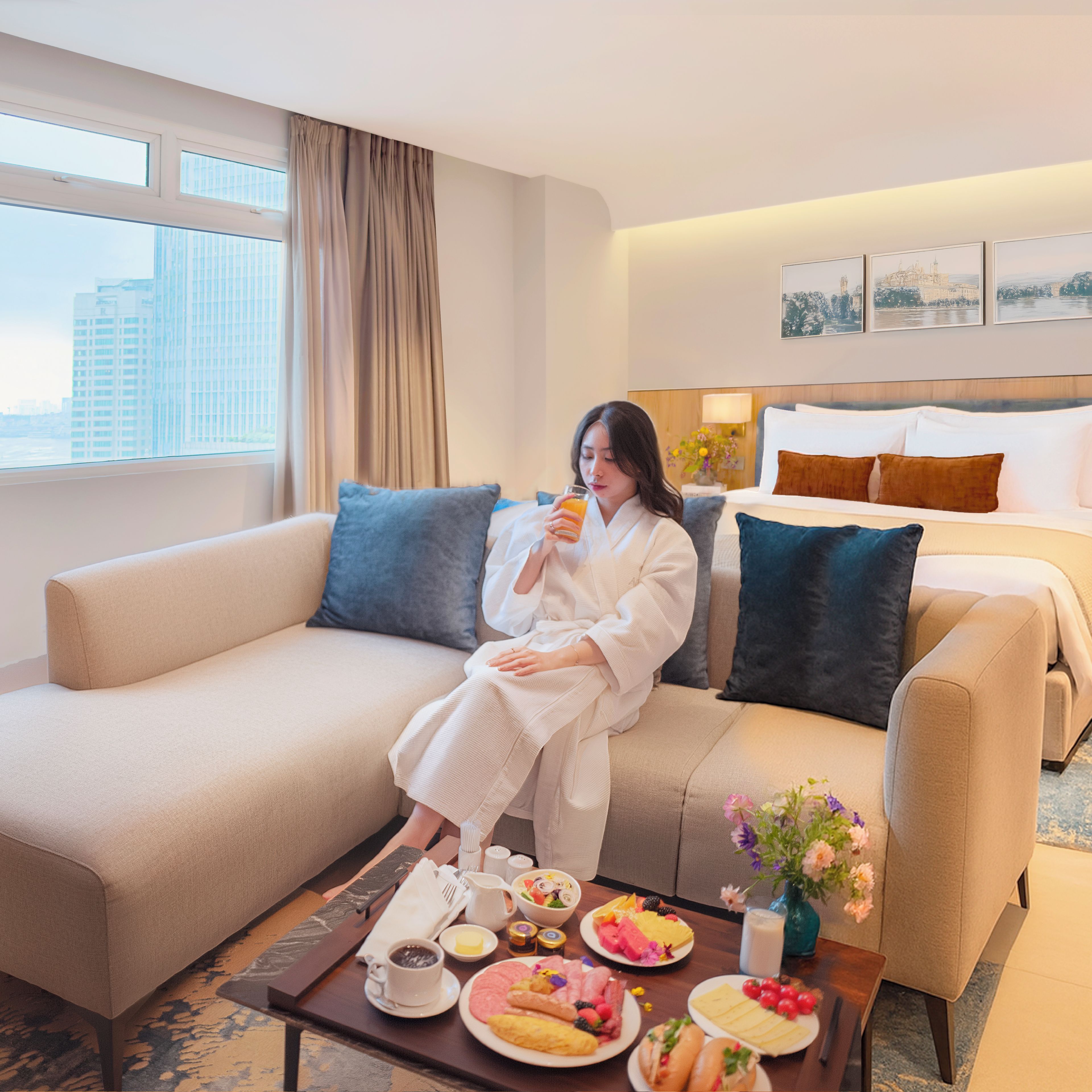 A hotel room with a sofa, a dining table with breakfast, and a bed with pillows.