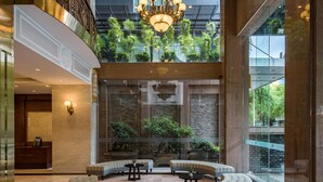 Un hall d’entrée spacieux avec un plafond haut, un lustre, un comptoir de réception et un espace détente offrant une vue sur un jardin intérieur luxuriant.