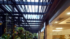 Un espace patio intérieur doté d’un plancher en bois, de chaises et de tables en métal, entouré de plantes en pot et d’un mur de verre.