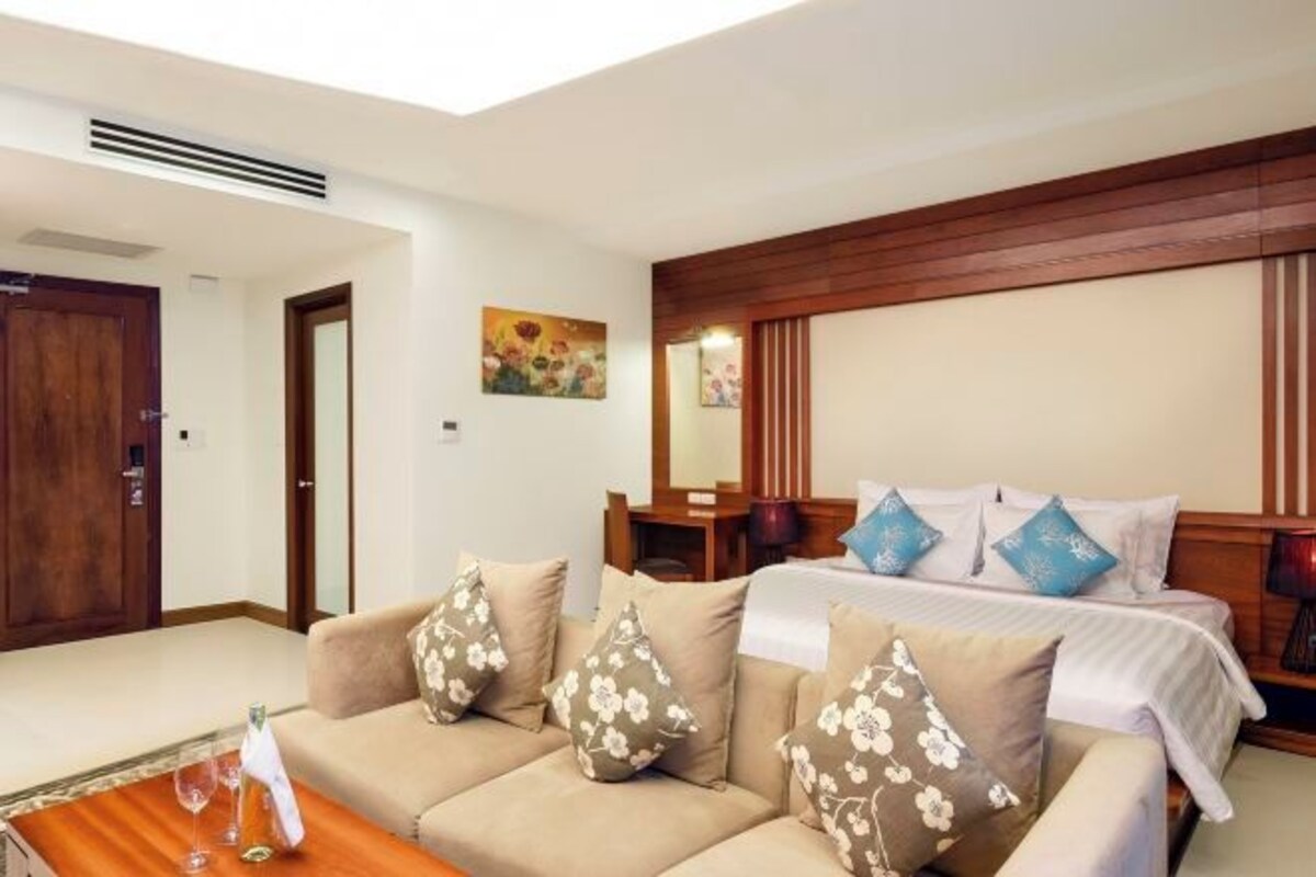 A hotel room with a bed, a sofa, a wooden headboard, a painting on the wall, and a door.