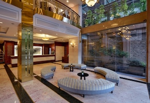 A spacious lobby with a high ceiling, a central seating area with sofas and a round table, and a large glass wall offering a view of indoor greenery.