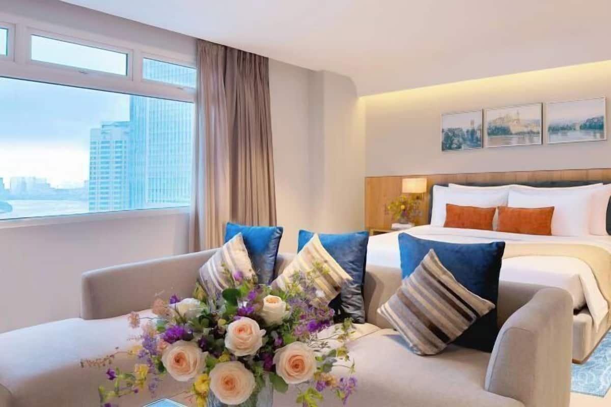 A hotel room with a bed, a sofa, a window with a city view, and a decorative flower arrangement.