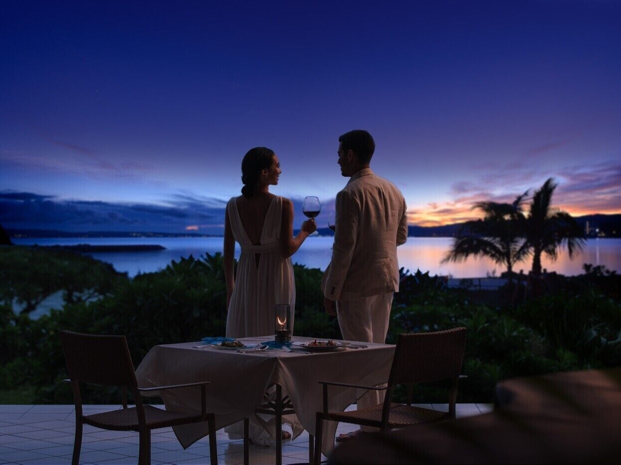 A couple dining at an outdoor table with a view of the ocean and palm trees at dusk.