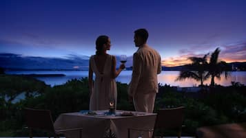 A couple dining at an outdoor table with a view of the ocean and palm trees at dusk.