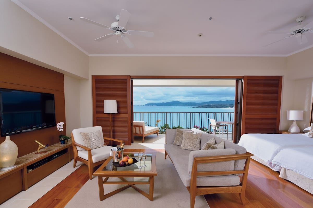 Ein modernes Hotelzimmer mit Balkon, Fernseher, Sofa, einem Couchtisch mit Obst und einem Bett.
