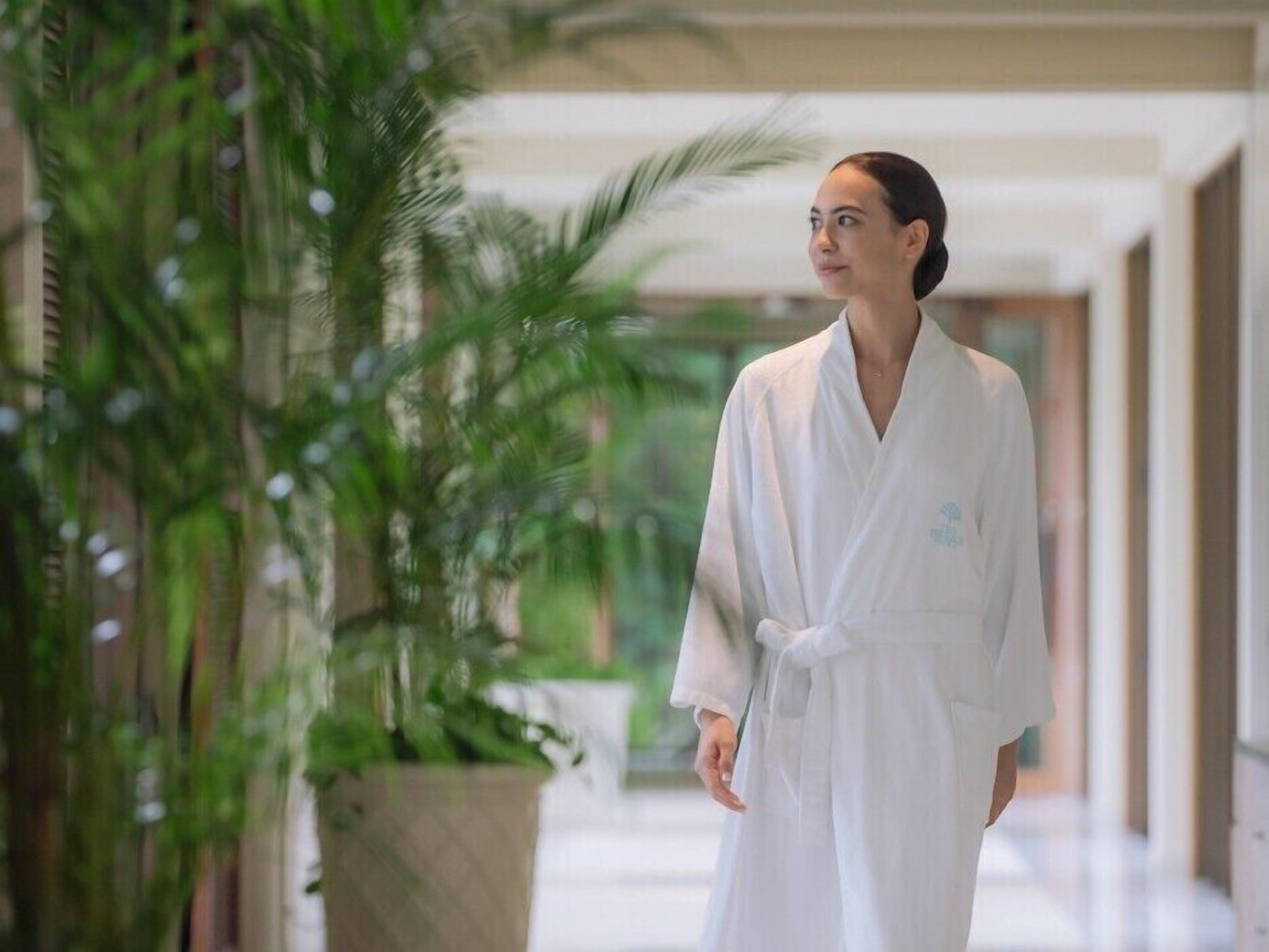 A person in a white robe standing in a hallway with a large potted plant and a view of a building outside.