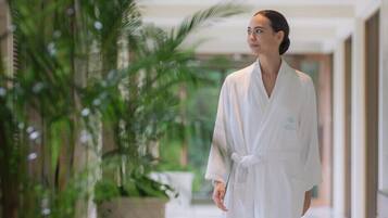 A person in a white robe standing in a hallway with a large potted plant and a view of a building outside.