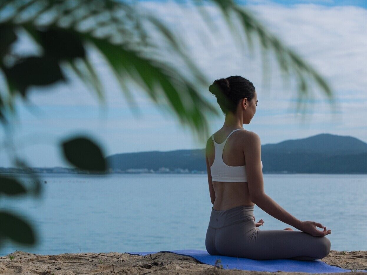Una persona meditando en una playa con vista al mar y montañas al fondo.