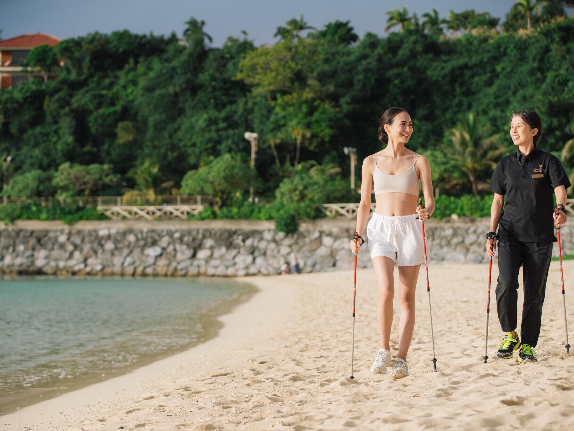 Two people walking on a sandy beach with a stone wall and greenery in the background.