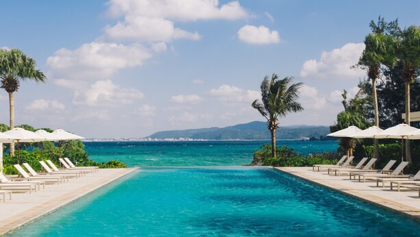 A pool with lounge chairs and umbrellas, surrounded by palm trees, with a view of the sea and distant mountains. - The Terrace Club Wellness Thalasso At Busena (Nago)