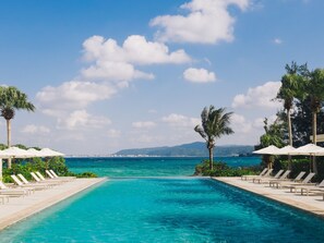 Une piscine bordée de chaises longues et de parasols, entourée de palmiers, et offrant une vue sur la mer et les montagnes lointaines.