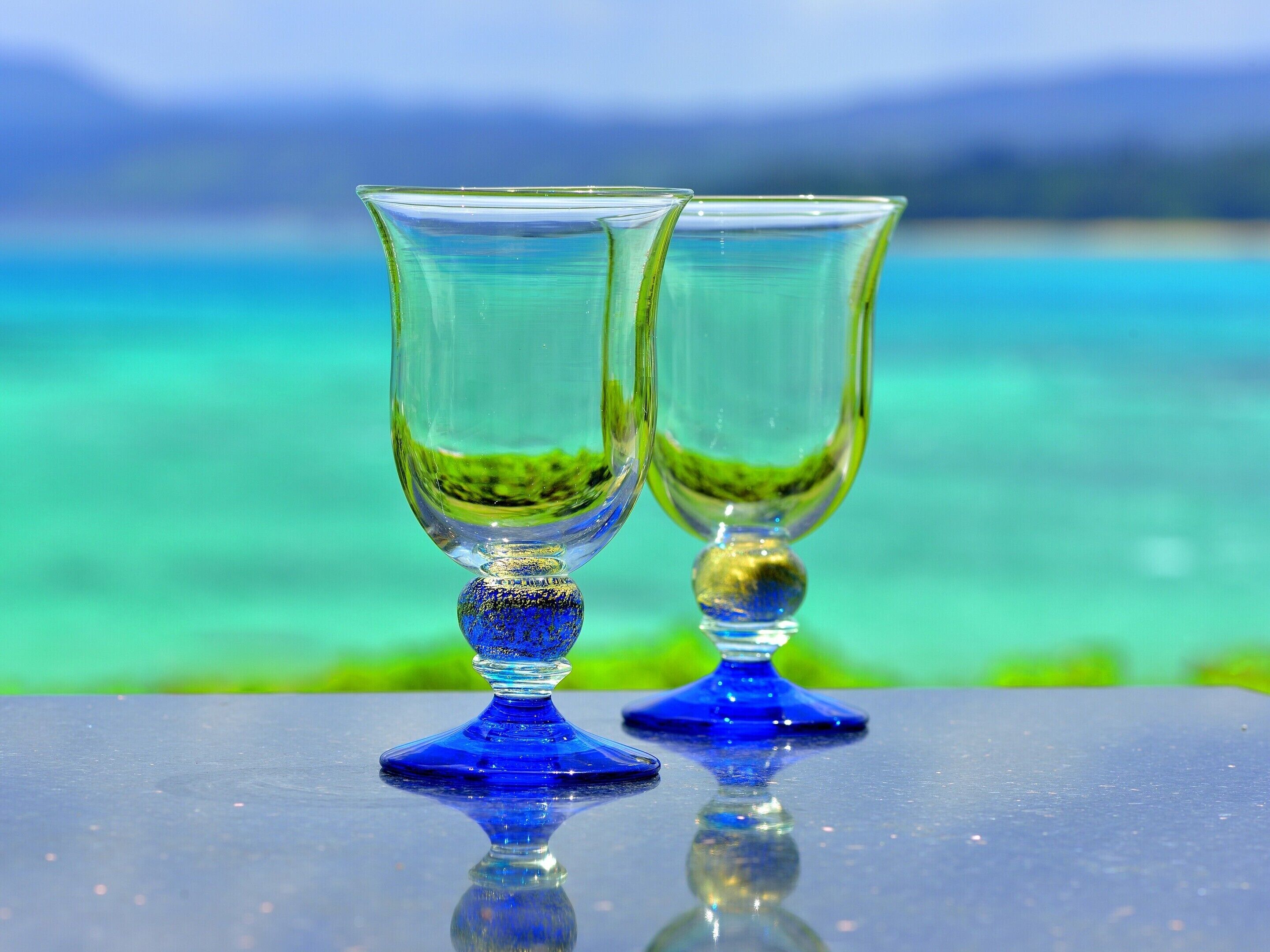 Dos copas de vidrio transparentes sobre una superficie reflectante, con un mar turquesa y montañas distantes al fondo.