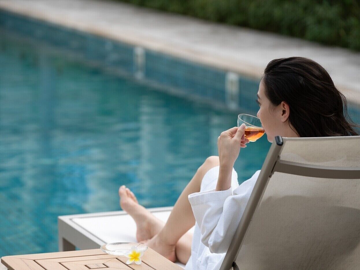 A person relaxing by a poolside lounge chair, holding a glass of a beverage.
