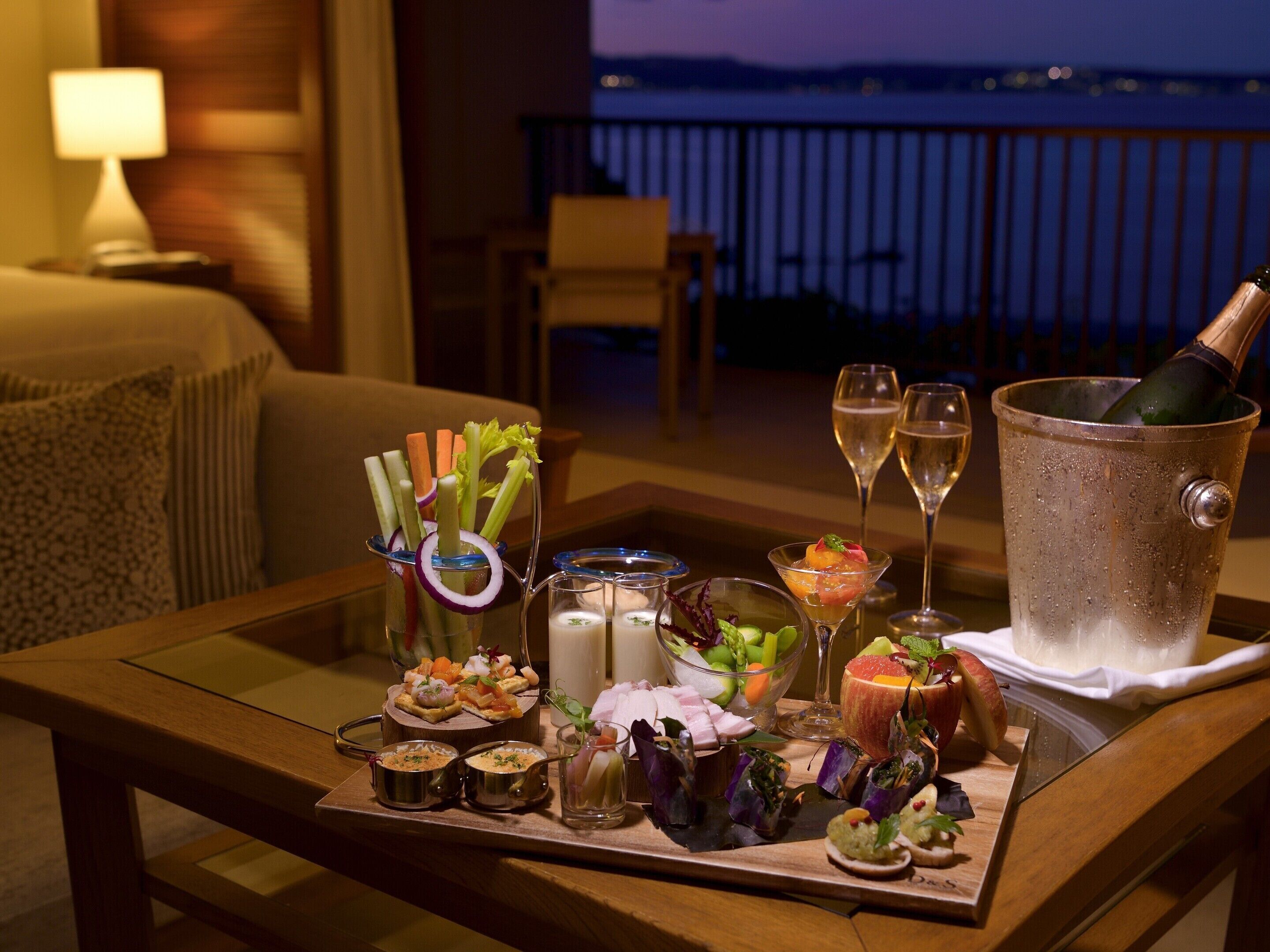 A table with a variety of appetizers and drinks, including a bottle of champagne, on a balcony overlooking a body of water.