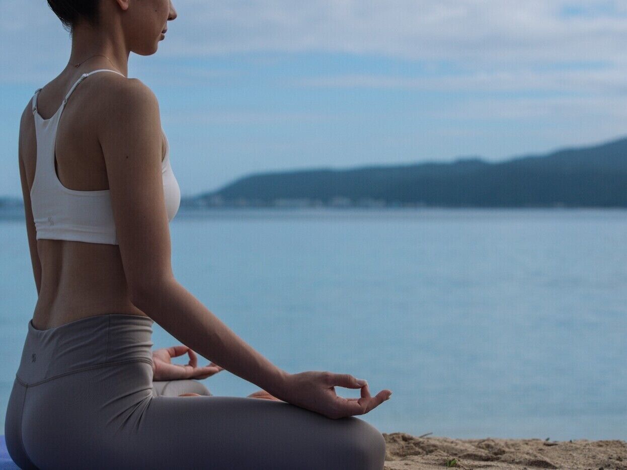 A person sitting in a meditative pose by a body of water with a mountainous backdrop.