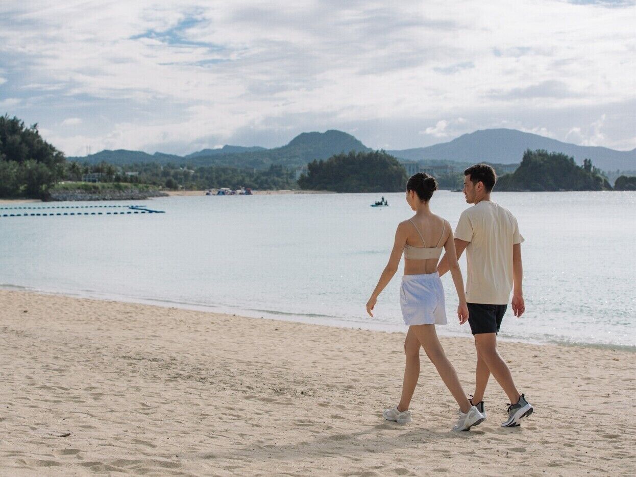 Una playa con una pareja caminando, un barco en el agua y montañas al fondo.