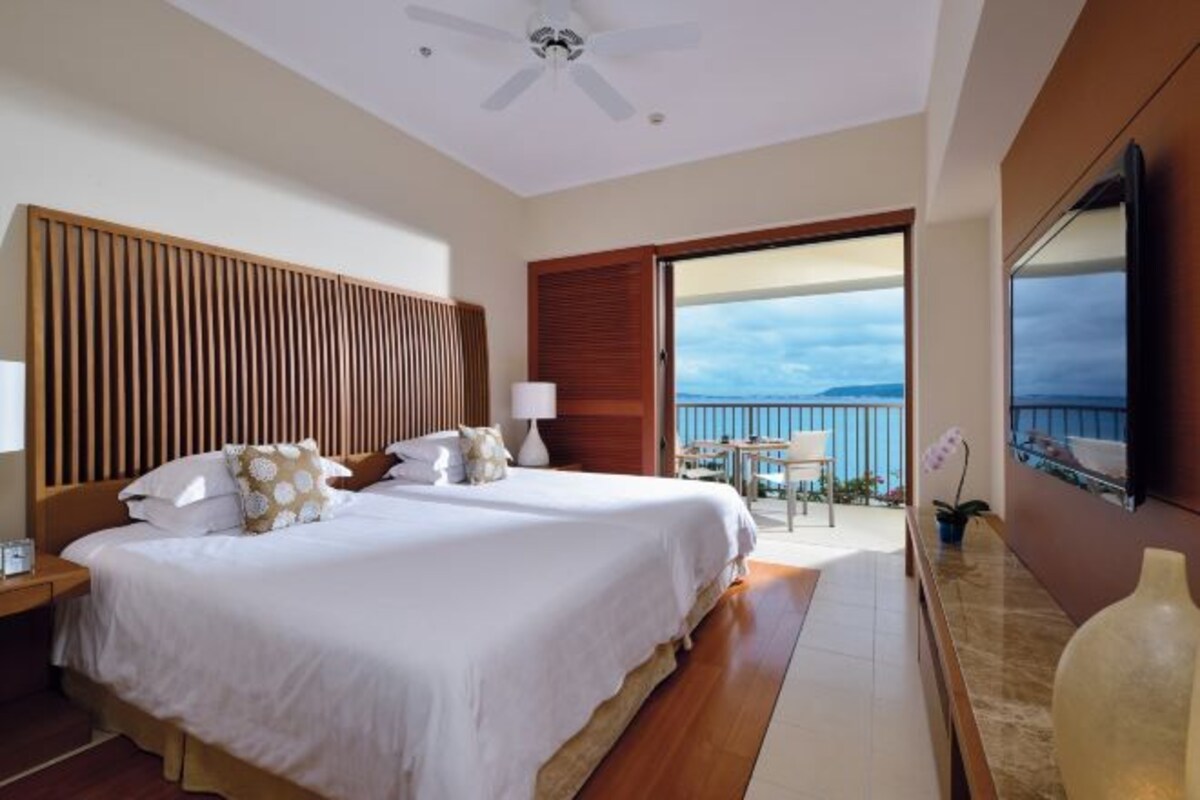 A hotel room with a large bed, a ceiling fan, a balcony with a view, and a TV on the wall.