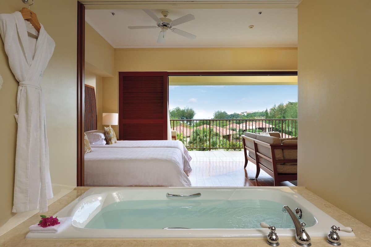 Ein Hotelzimmer mit einer großen Badewanne, zwei Betten, einem Balkon mit Blick und einem Deckenventilator.