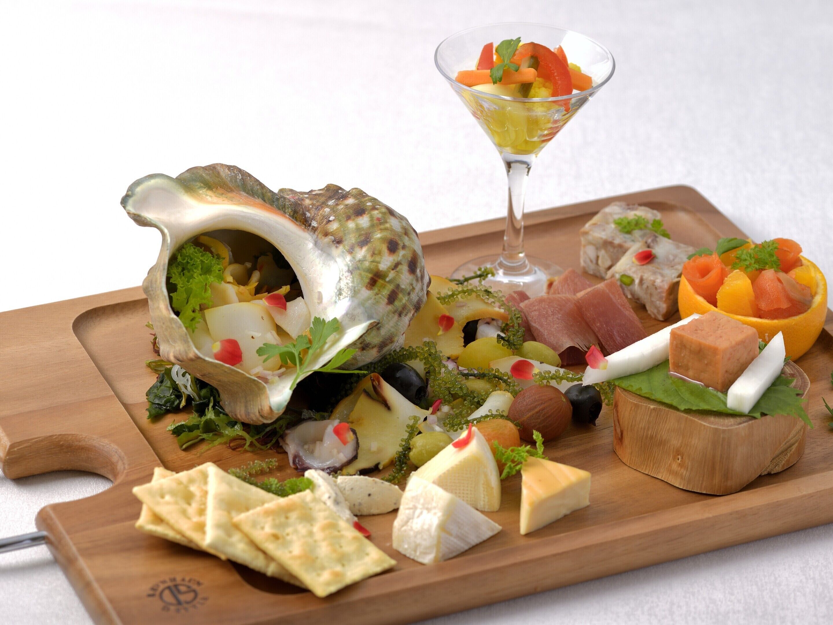 A wooden board with various cheeses, crackers, and a salad in a shell.